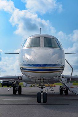 Giriş merdivenleri geniş apronla park edilmiş özel bir uçağın ön görüntüsü. Uçak dikey çerçevelenmiş, bulutlu gökyüzü ve parlak burun konisinde yansımaları var..