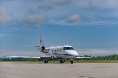 Geniş mavi gökyüzünün altında, uzak bulutlarla pistte kullanışlı jet taksiler. Ön dörtte bir açıyla yakalanan bileşim, hem motorları, hem iniş takımlarını ve uçak gövdesini gösteriyor. Resim kopya için negatif boşluk içeriyor.