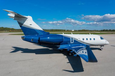 Yan görünüş, biraz yüksek açı, modern mavi ve beyaz yönetici jetinin tam fotoğrafı açık bulutlu mavi gökyüzünün altında bir havaalanı pistinde park edilmiş..