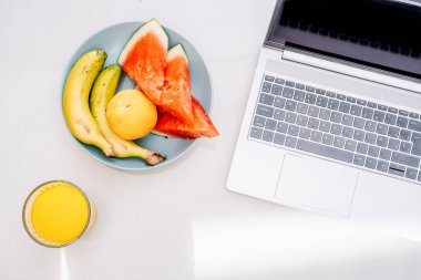 Taze meyve dilimleri ve meyve suyu beyaz bir masada açık bir dizüstü bilgisayarın yanında.