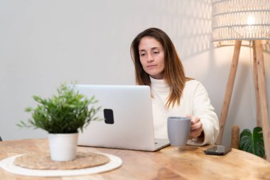 Ahşap masada dizüstü bilgisayar kullanan, rahat bir evde kahvenin keyfini çıkaran bir kadın.