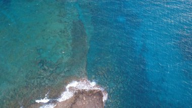 Gün batımında deniz kenarı, kayalık kıyılar ve sakin okyanusun yer aldığı manzaralı bir sahil şeridinin yüksek kaliteli hava fotoğrafı. Denize yansıyan altın saat ışığı, barışçıl ve atmosferik bir kıyı manzarası yaratır. Seyahat ve yaz tatili için ideal.