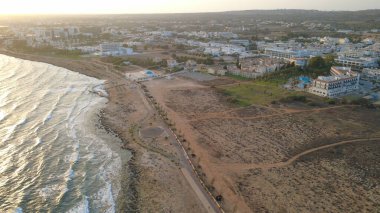 Gün batımında kayalık kıyı şeridi ve sakin denizi olan bir kıyı kasabasının yüksek kaliteli hava fotoğrafı. Güneş ışığı, okyanus suyunu, deniz kenarındaki binaları ve temiz gökyüzü altındaki manzaralı kıyı manzarasını yansıtıyor. Seyahat, turizm, tatil, yaz için ideal.