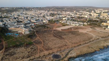Gün batımında kayalık kıyı şeridi ve sakin denizi olan bir kıyı kasabasının yüksek kaliteli hava fotoğrafı. Güneş ışığı, okyanus suyunu, deniz kenarındaki binaları ve temiz gökyüzü altındaki manzaralı kıyı manzarasını yansıtıyor. Seyahat, turizm, tatil, yaz için ideal.