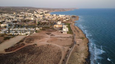 Gün batımında kayalık kıyı şeridi ve sakin denizi olan bir kıyı kasabasının yüksek kaliteli hava fotoğrafı. Güneş ışığı, okyanus suyunu, deniz kenarındaki binaları ve temiz gökyüzü altındaki manzaralı kıyı manzarasını yansıtıyor. Seyahat, turizm, tatil, yaz için ideal.