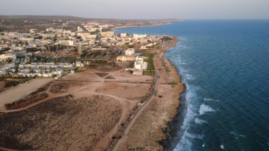 Gün batımında kayalık kıyı şeridi ve sakin denizi olan bir kıyı kasabasının yüksek kaliteli hava fotoğrafı. Güneş ışığı, okyanus suyunu, deniz kenarındaki binaları ve temiz gökyüzü altındaki manzaralı kıyı manzarasını yansıtıyor. Seyahat, turizm, tatil, yaz için ideal.