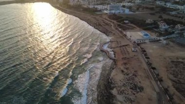 Altın saat boyunca kayalık kıyı şeridi ve okyanus dalgaları olan bir sahil kasabasının sinematik hava aracı görüntüleri. Sakin deniz, sahil yolu ve açık gökyüzünün altında sahil boyunca binalar. Seyahat, turizm, yaz tatili, gayrimenkul, doğa için ideal.