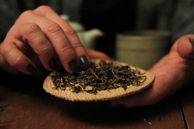 Sıcak ışıkta seramik çaydanlık, fincanlar ve ahşap bir masada gevşek yaprak çayıyla samimi çay seremonisi sahnesi. Yavaş yaşam, farkındalık, konfor, ev ritüelleri ve çay kültürü kavramlarına uygundur..