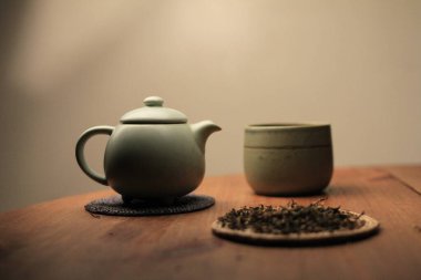 Sıcak ışıkta seramik çaydanlık, fincanlar ve ahşap bir masada gevşek yaprak çayıyla samimi çay seremonisi sahnesi. Yavaş yaşam, farkındalık, konfor, ev ritüelleri ve çay kültürü kavramlarına uygundur..