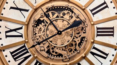 A large, open-faced wall clock with a distressed gold-wash metal finish and exposed, moving 3D gears. Close up of elegant Antique clock featuring roman numerals, classic design