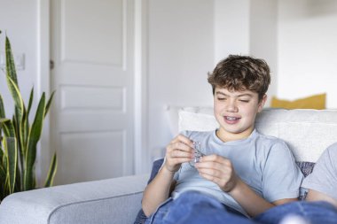 Diş telleriyle kanepede oturan, metal bulmacaları çözen, evde boş vakitlerin tadını çıkaran genç bir çocuk.