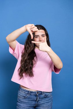 Elleriyle bir parmak çerçevesi oluştururken gülümseyen genç bir kadın, mavi bir arka plan karşısında perspektif buluyor, kot pantolon ve pembe bir tişört giyiyor..