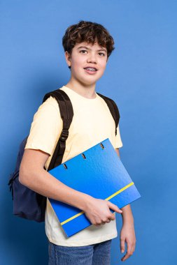 Teen boy with braces ready for school, wearing backpack , smiling on blue background