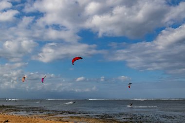 Renkli uçurtmacılar parlak mavi bir gökyüzünün altında Bali 'nin sığ kıyı sularında süzülüyorlar. Enerjik hareket, dramatik bulutlar ve tropikal manzara seyahat ve spor görüntüleme için mükemmel bir açık hava macera sahnesi yaratır..