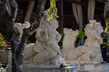 Yeşillik bir tropikal Bali bahçesine yerleştirilmiş süslü taş heykeller. Ayrıntılı heykellerde, seyahat, miras ve mimari temalar için ideal huzurlu ve kültürel bir atmosfer yaratan geleneksel Balili sanatsal motifler yer alıyor..