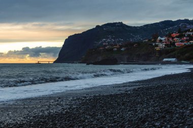 Portekiz 'in Madeira şehrinde kayalık bir çakıl taşı plajında, bulutlu bir akşamın gölgesinde gölgelendirilmiş, Atlantik' in güçlü dalgaları, altın ufuk ışığı ve karanlık uçurumları ile Madeiradramatik günbatımında Huysuz Gün Batımı..