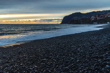 Portekiz 'in Madeira şehrinde kayalık bir çakıl taşı plajında, bulutlu bir akşamın gölgesinde gölgelendirilmiş, Atlantik' in güçlü dalgaları, altın ufuk ışığı ve karanlık uçurumları ile Madeiradramatik günbatımında Huysuz Gün Batımı..