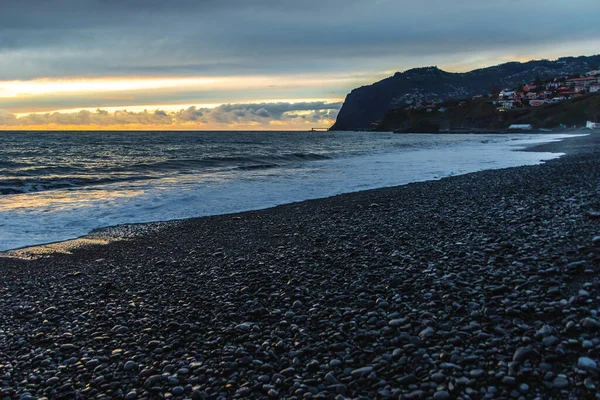 Portekiz 'in Madeira şehrinde kayalık bir çakıl taşı plajında, bulutlu bir akşamın gölgesinde gölgelendirilmiş, Atlantik' in güçlü dalgaları, altın ufuk ışığı ve karanlık uçurumları ile Madeiradramatik günbatımında Huysuz Gün Batımı..