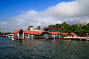 Renkli tekneler Livingston, Guatemala 'daki Karayipler rıhtımına yanaştı. Tropikal palmiye ağaçları, canlı binalar ve dramatik bulutlar canlı bir sahil atmosferi yaratıyor..