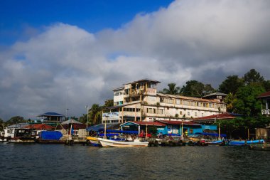 Renkli tekneler Livingston, Guatemala 'daki Karayipler rıhtımına yanaştı. Tropikal palmiye ağaçları, canlı binalar ve dramatik bulutlar canlı bir sahil atmosferi yaratıyor..
