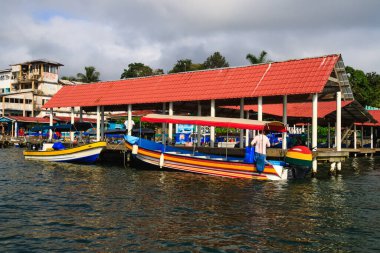 Renkli tekneler Livingston, Guatemala 'daki Karayipler rıhtımına yanaştı. Tropikal palmiye ağaçları, canlı binalar ve dramatik bulutlar canlı bir sahil atmosferi yaratıyor..