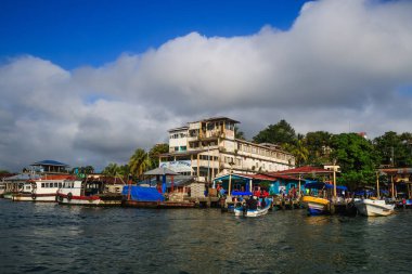 Renkli tekneler Livingston, Guatemala 'daki Karayipler rıhtımına yanaştı. Tropikal palmiye ağaçları, canlı binalar ve dramatik bulutlar canlı bir sahil atmosferi yaratıyor..