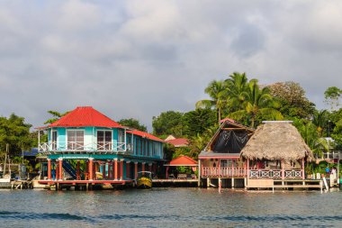 Renkli tekneler Livingston, Guatemala 'daki Karayipler rıhtımına yanaştı. Tropikal palmiye ağaçları, canlı binalar ve dramatik bulutlar canlı bir sahil atmosferi yaratıyor..