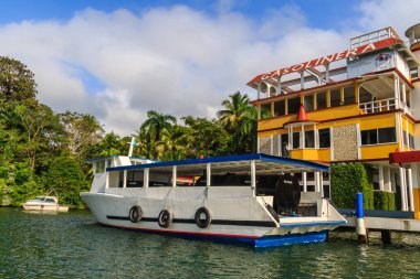 Renkli tekneler Livingston, Guatemala 'daki Karayipler rıhtımına yanaştı. Tropikal palmiye ağaçları, canlı binalar ve dramatik bulutlar canlı bir sahil atmosferi yaratıyor..