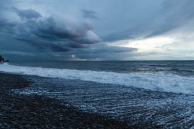 Atlantik dalgaları alacakaranlıkta karanlık bir çakıl taşı plajına çarptığında Madeira 'nın engebeli volkanik kıyı şeridinde dramatik fırtına bulutları toplanıyor..