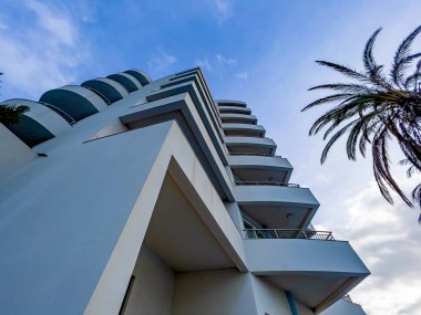 Mavi gökyüzü altında kıyı yaşamını, seyahatini, yaz tatilini ve çağdaş mimari tasarımı çağrıştıran kıvrımlı balkonları ve palmiyeleri olan modern beyaz bir apartmanın alçak açılı görüntüsü..