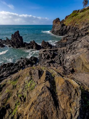 Portekiz, Madeira 'daki Atlantik Okyanusu boyunca dramatik kayalık kayalıklar. Parlak, yansıtıcı bir gökyüzü altında engebeli volkanik oluşumlara çarpan dalgalar..