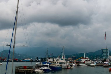 Şiddetli bulutların altında birden fazla rıhtımlı teknesi olan fırtınalı bir liman limanı, kıyı kenti silueti ve uzak dağlar karamsar hava, seyahat, balıkçılık ve deniz atmosferini iletiyor. Batumi, Georgia