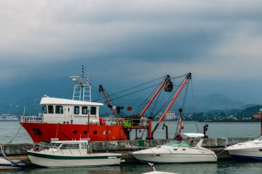 Beyaz zevk teknelerinin yanındaki beton iskeleye demirlemiş kırmızı bir balıkçı teknesi sisli dağlara karşı kurulmuş ve bulutlu bir gökyüzü sahil sanayisini, sakin liman hayatını ve kasvetli havayı aktarıyor. Batumi, Georgia