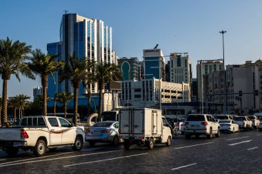 Yoğun şehir trafiği, Doha 'nın altın saatinde modern cam gökdelenlerin önündeki palmiye çizgili bulvar boyunca ilerlerken, şehir içi ulaşımı, çağdaş mimari ve kıyı metropolünde sıcak bir akşam ışığı vurgulanıyor..
