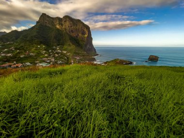 Portekiz 'in Madeira Adası' nın kuzey kıyısındaki Porto da Cruz köyünün manzaralı manzarası Atlantik Okyanusu üzerinde yükselen dramatik Penha dguia volkanik uçurumu ve kıyı kasabasını çevreleyen yemyeşil tepeler..