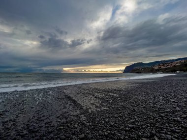 Güçlü Atlantik dalgaları, Portekiz 'in engebeli Madeira kıyı şeridinde, arka planda dramatik bulutlar ve sahil kayalıkları bulunan karanlık bir volkanik çakıl taşını kıyıya vuruyor..