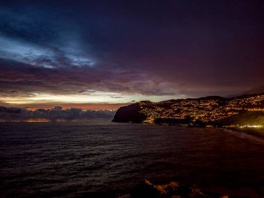 Alacakaranlıkta Madeira 'nın engebeli uçurumları boyunca bir sahil kasabası ışıldıyor, sakin okyanusta yansıyan sıcak sokak ışıkları dramatik, renkli bir gökyüzü ve ufukta görünen uzak bulut çizgisi altında..