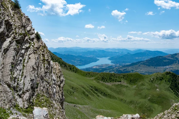 Kayalık kayalıkları, yeşil vadileri ve bulutlu mavi gökyüzünün altında uzak bir gölü olan manzaralı dağ manzarası..