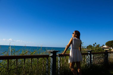 Korkuluklara yaslanan Kadın Güneşli bir balkonda Denize Bakıyor