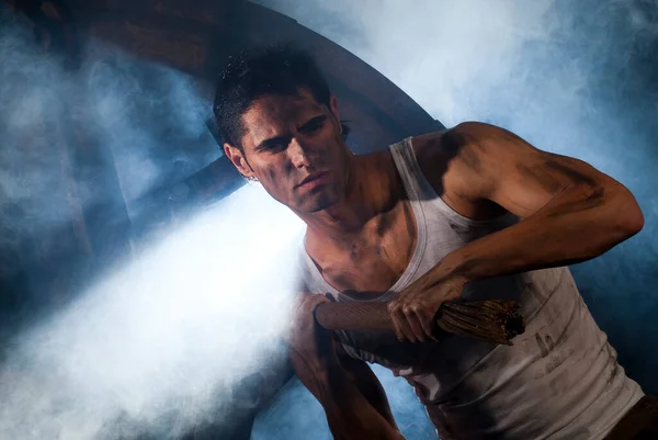 Kirli yüzü ve kolları olan güçlü bir adam fiziksel çaba sarf ediyor, tozlu, karanlık ve yoğun bir ortamda bir alet tutuyor.