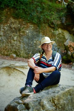 Şapkalı, gülümseyen orta yaşlı bir kadın doğal ortamdaki bir kayanın üzerinde oturuyor ve dinleniyor, Urederra, Baquedano, Navarra, İspanya, Avrupa