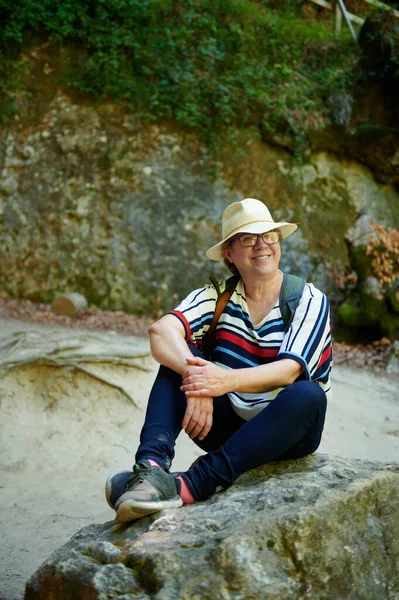 Şapkalı, gülümseyen orta yaşlı bir kadın doğal ortamdaki bir kayanın üzerinde oturuyor ve dinleniyor, Urederra, Baquedano, Navarra, İspanya, Avrupa