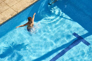 Çocuk yüzme havuzuna atlıyor, su sıçratıyor. Güneş mavi fayanslı suyun üzerinde parlar, yazı ve boş zamanı yansıtır.
