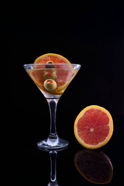 Portakal dilimi ve zeytinlerle süslenmiş, koyu yansıtıcı bir yüzeyde martini bardağında canlandırıcı bir kokteyl.