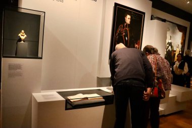 Bu fotoğraf, bir müze ziyaretçisinin klasik bir portrenin altındaki vitrini incelemesini gösteriyor. Bu etkileşim sanat eseriyle izleyici arasında bir diyalog yaratır. Saray müzesinin eğitici atmosferini yansıtıyor..