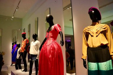 Fotoğraf modern giyim tarzında giyinmiş bir grup mankeni gösteriyor. Bir manken dikkat çekici bir kırmızı gece elbisesi giyerken, diğerleri çeşitli çağdaş giysiler giyer..