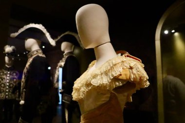 Antik bir elbise giyen bir kadın mankenin yakın çekimi. Arka planda tarihi giysiler giyen erkek mankenler görülüyor..