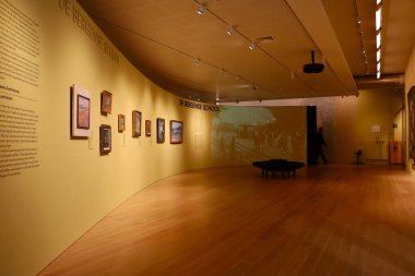 Sıcak sarı tonlu boş bir müze salonunda duvarlar boyunca sergilenen tablolar ve merkezde orijinal bir oturma tezgahı yer alıyor. Ziyaretçilerin yokluğunda sükunet ve mekansal tasarım vurgulanıyor.