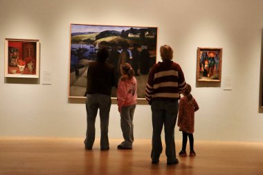 Aralarında iki yetişkin kadın ve iki çocuğun da bulunduğu dört kişi, bir tabloyu incelerken bir müze salonunda yan yana dururlar. Sahne, sanatla merak ve eğitimsel bağlılık paylaşıyor. Bu resim aile müzesi ziyaretlerini temsil ediyor.