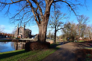 Erken bir bahar parkının ön planında yapraksız bir ağaç duruyor. Arkasında, büyük tarihi bir binaya açılan bir köprü var..